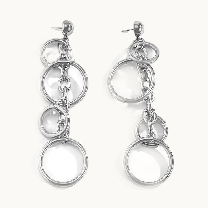 Une paire de boucles d'oreilles est exposée devant un fond beige.  Ce sont des boucles d'oreille pendant chaîne sur laquelle est accrochée des anneaux de différentes tailles. Elles sont en argent. 