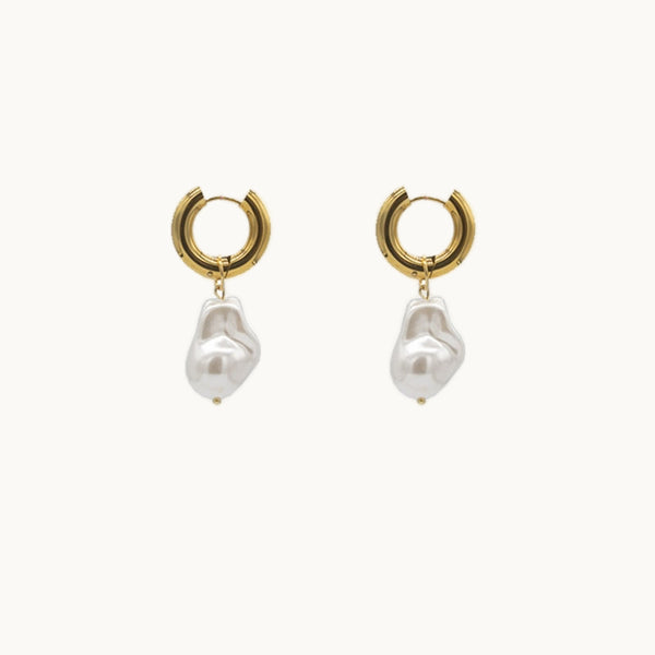 Une paire de boucles d'oreilles est exposée devant un fond beige.  Ce sont des créoles dorées petites et épaisses sur lesquelles pendent un perle. 