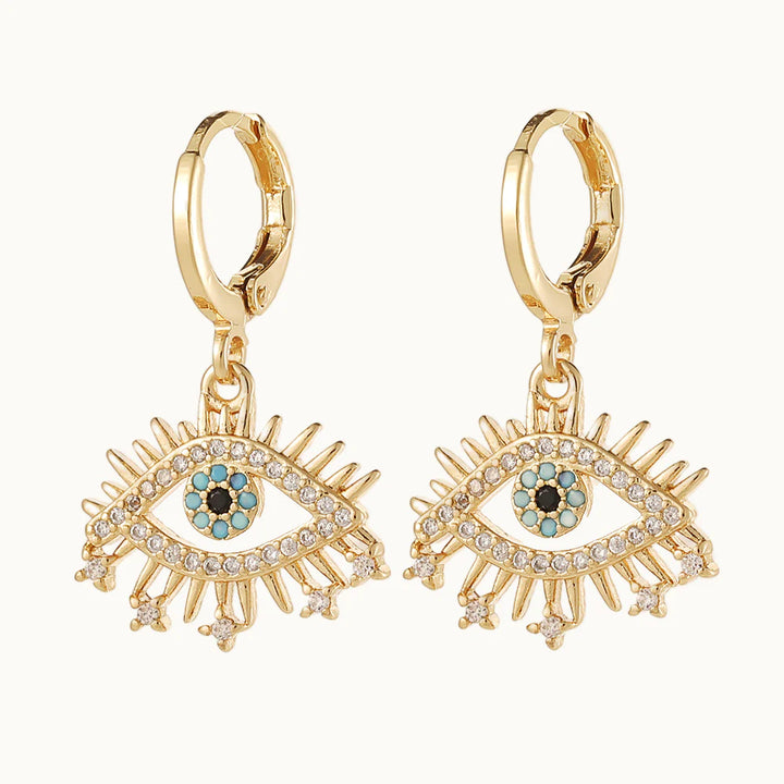 Une paire de boucles d'oreilles est exposée devant un fond beige.Ce sont des boucles d'oreille en forme d'anneau sur lequel pend un oeil doré. L'iris et la pupille sont ornés de strass bleu et noir. Elles sont en dorées et l'oeil et les cils sont ornées de strass. 