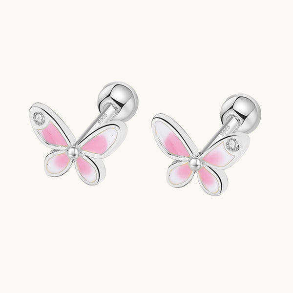 Une paire de boucles d'oreilles est exposée devant un fond beige.  Ce sont des papillons en argent avec des ailes roses et blanches. Il y a un strass sur l'une des ailes. 