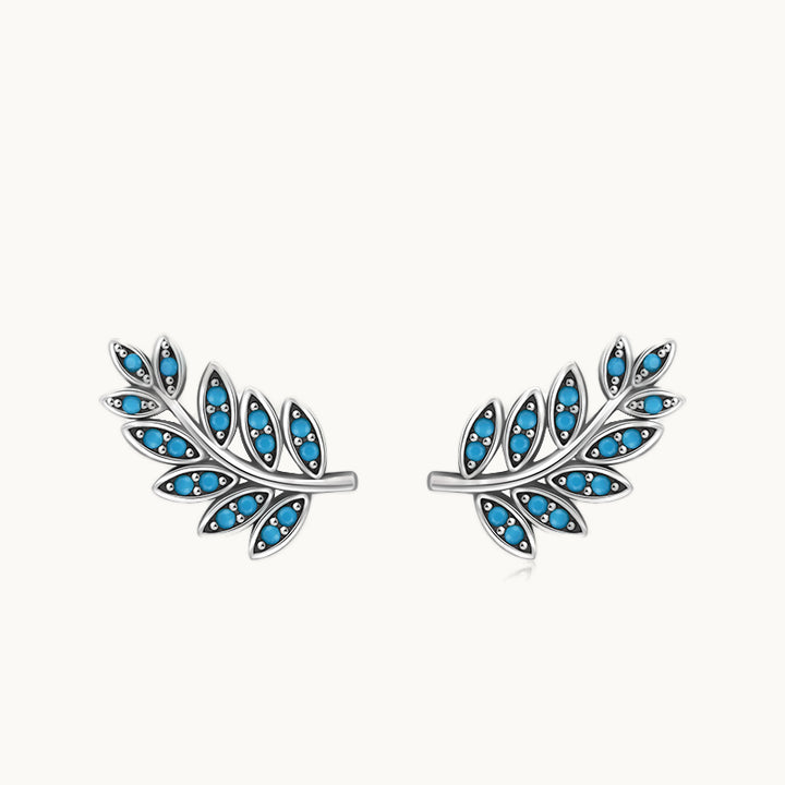 Une paire de boucles d'oreilles est exposée devant un fond beige.  Ce sont des boucles d'oreille grimpantes en forme de feuilles.  Elles sont en argent 925 et sont ornées de pierres turquoises.