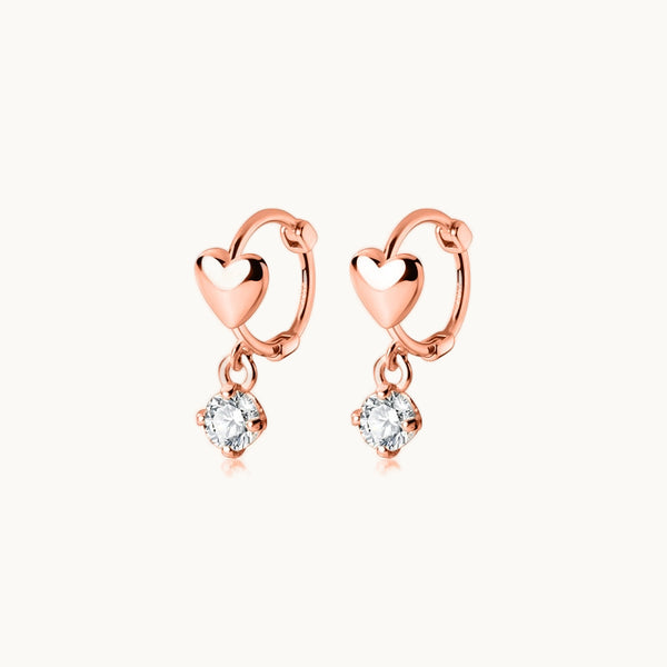 Une paire de boucles d'oreilles est exposée devant un fond beige.  Ce sont des boucles d'oreille dormeuses ornées d'un coeur et d'un diamant pendant. Elles sont en argent 925 plaqué or rose.