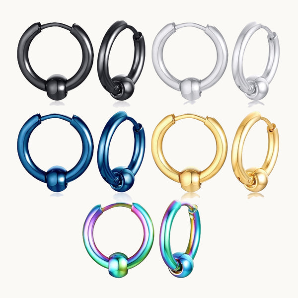 Cinq paires de boucles d'oreilles sont exposées devant un fond beige.  Ce sont des anneaux de différentes couleurs en acier sur lesquelles il y a une boule métallique. 