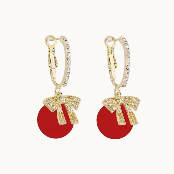 Une paire de boucles d'oreille est exposée devant un fond beige.  Ce sont des boucles d'oreille dormeuses. L'attache est ornée de strass sur le devant. Pendent de cette attache un noeud doré orné de strass et une boule de noël rouge. Les boucles sont dorées.