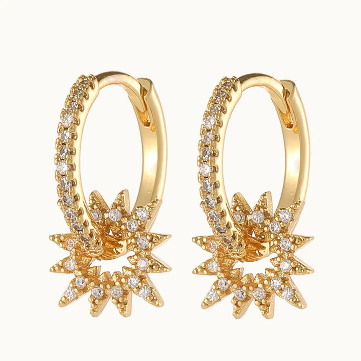 Une paire de boucles d'oreilles est exposée devant un fond beige. Ce sont des boucles d'oreille en forme d'anneau sur lequel pend un soleil ajouré. La boucle d'oreille est ornée de strass. Elles sont en dorées. 