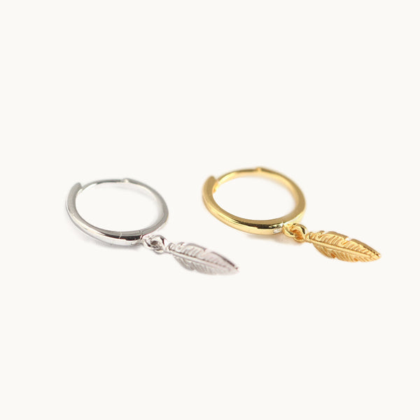 Des boucles d'oreilles sont exposées devant un fond beige.  Ce sont des boucles pendantes composées d'un anneau sur lequel pend une plume. Elles sont en argent 925 et argent 925 plaqué or. 