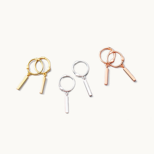 Trois paires de boucles d'oreilles sont exposées devant un fond beige.  Ce sont des boucles d'oreille composées d'un anneau sur lequel pend une pièce rectangulaire longue. Ce sont de petites créoles avec un long rectangle en relief qui pend dessus. Il y a une paire en argent 925, une en argent 925 plaqué or et une en argent 925 plaqué or rose