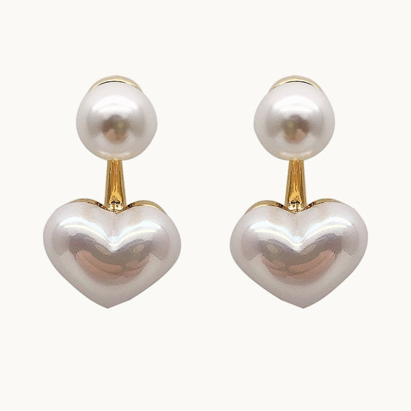Une paire de boucles d'oreilles est exposée devant un fond beige.  Ce sont des boucles d'oreilles avec une perle ronde, pendante dorée avec un coeur en perle sur le bas. 