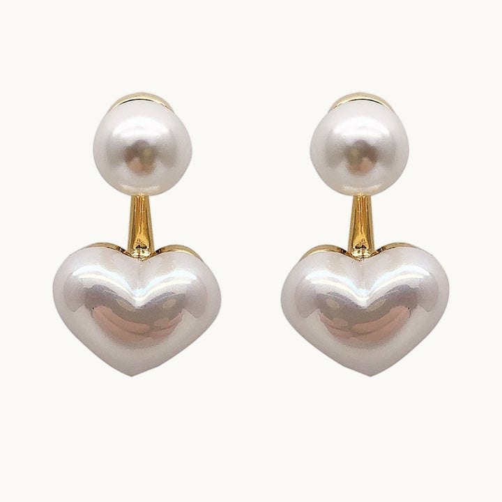 Une paire de boucles d'oreilles est exposée devant un fond beige.  Ce sont des boucles d'oreilles avec une perle ronde, pendante dorée avec un coeur en perle sur le bas. 