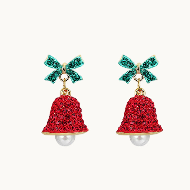 Une paire de boucles d'oreille est exposée devant un fond beige.  Ce sont des boucles d'oreille en forme de cloche pendante. Elles sont ornées de strass. Il y a un noeud vert au-dessus de la cloche rouge. Dans la cloche il y a une perle. Elles sont dorées.