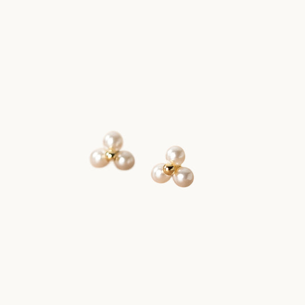 Une paire de boucles d'oreilles est exposée devant un fond beige.  Ce sont des boucles fines avec une perle métallique en argent plaqué or au centre entourée par trois perles artificielles. 