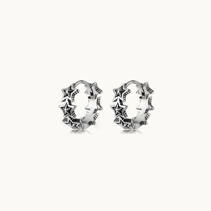 Une paire de boucles d'oreilles est exposée devant un fond beige.  Ce sont des créoles en forme d'étoiles. Elles sont en argent 925 avec un effet vieilli. 