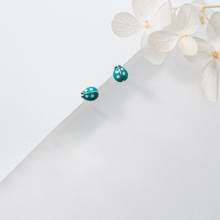 Boucle d'oreille coccinelle bleue en argent 925 - Deux coccinelles sur une feuille de papier avec des fleurs blanches.