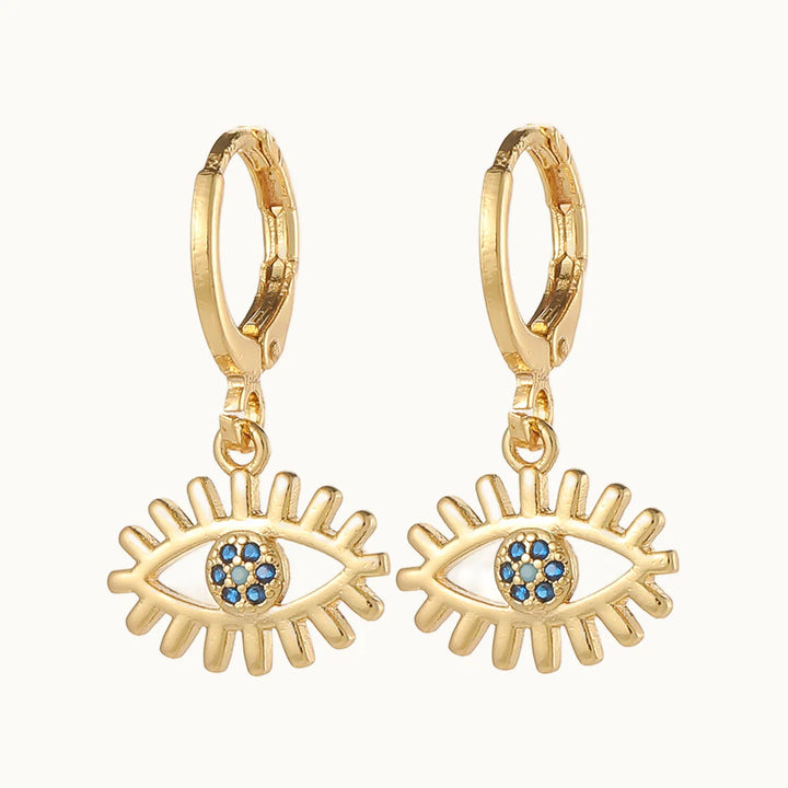 Une paire de boucles d'oreilles est exposée devant un fond beige.  Ce sont des boucles d'oreille en forme d'anneau sur lequel pend un oeil doré. L'iris et la pupille sont ornés de strass bleu. Elles sont en dorées. 