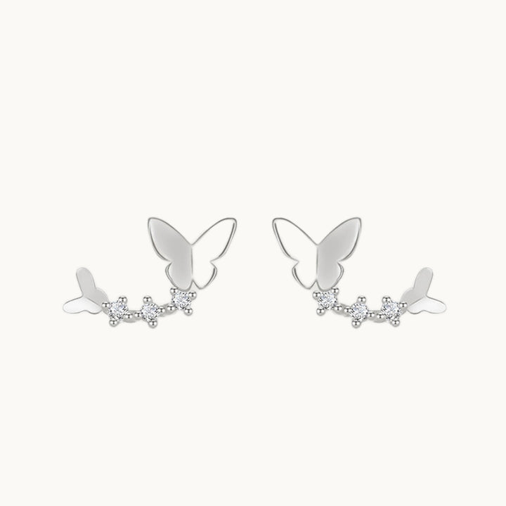 Une paire de boucles d'oreilles est exposée devant un fond beige.  Ce sont des boucles grimpantes constituées d'un papillon à chaque extrémité avec des strass. Elles sont en argent 925