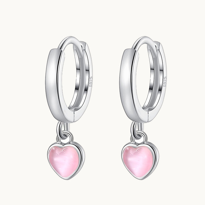 Une paire de boucles d'oreilles est exposée devant un fond beige.  Ce sont des anneaux épais en argent 925 sur lesquels pend un coeur comme une pierre d'opale colorée. Le coeur est de couleur rose. 