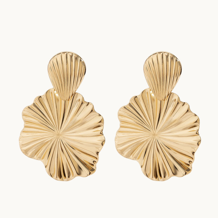 Une paire de boucles d'oreille est exposée devant un fond beige.  Ce sont des boucles d'oreille pendante. Le métal est doré et plissé. Elles sont composées d'un coquillage d'où pend une fleur. 