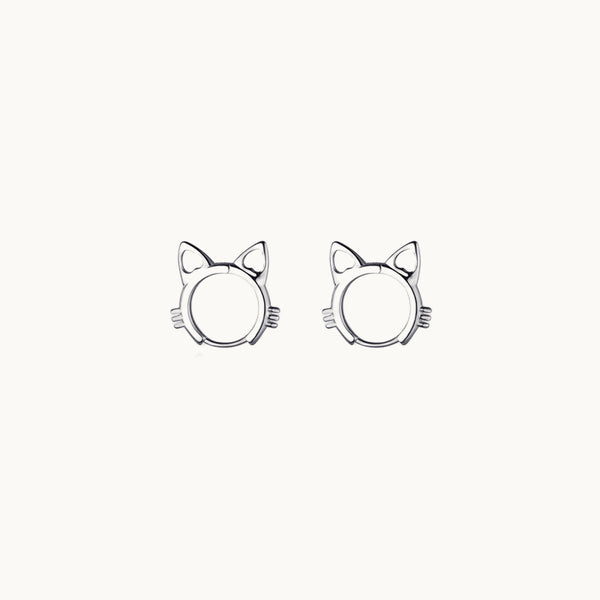 Une paire de boucles d'oreilles est exposée devant un fond beige.  Ce sont des anneaux en forme de tête de chat.  Ils sont en argent. 