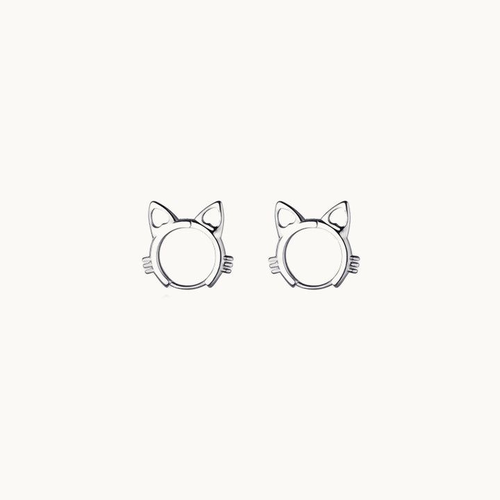 Une paire de boucles d'oreilles est exposée devant un fond beige.  Ce sont des anneaux en forme de tête de chat.  Ils sont en argent. 