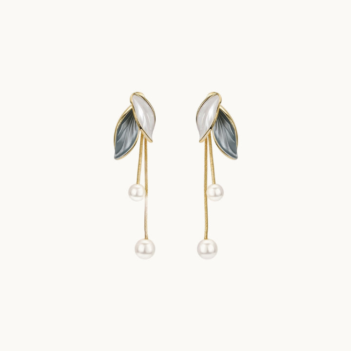 Une paire de boucles d'oreille est exposée devant un fond beige.  Ce sont des boucles d'oreille en forme de feuilles. Les feuilles sont en strass, l'une blanche et l'autre gris vert. De ces feuilles pendent deux chaines en maille serpent au bout desquelles une perle. 