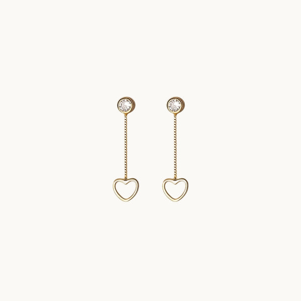 Une paire de boucles d'oreilles est exposée devant un fond beige.  Ce sont des boucles composées d'un diamant de zirconium rond sur lequel pend une chaîne. Au bout de la chaîne il y a un petit coeur. Elle sont en argent 925 plaqué or.