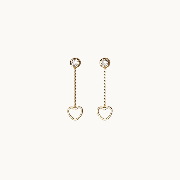 Une paire de boucles d'oreilles est exposée devant un fond beige.  Ce sont des boucles composées d'un diamant de zirconium rond sur lequel pend une chaîne. Au bout de la chaîne il y a un petit coeur. Elle sont en argent 925 plaqué or.