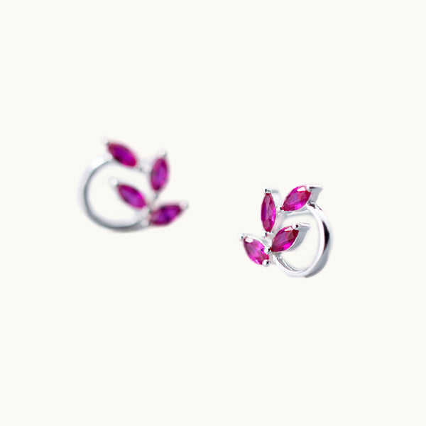 Une paire de boucles d'oreilles est exposée devant un fond beige.   Ce sont des boucles d'oreille rondes ornées de feuilles en diamants colorés. Elles sont en argent 925 et les feuilles sont de couleur rose. 
