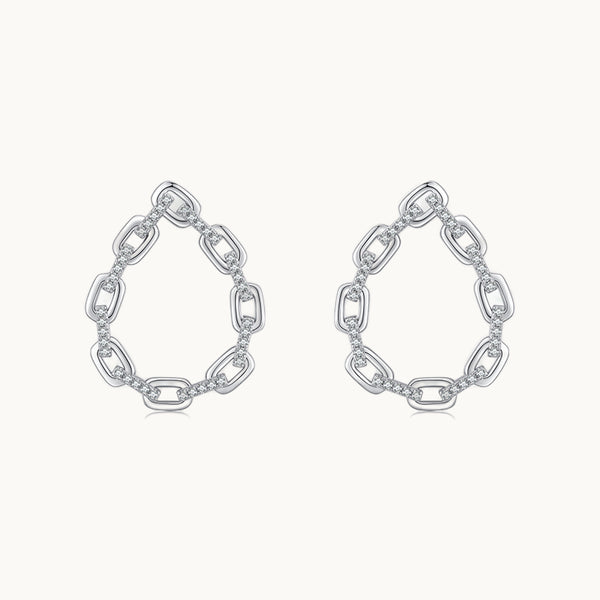 Une paire de boucles d'oreilles est exposée devant un fond beige.  Ce sont des boucles d'oreille en forme de goutte. Elles sont constituées de maillons de chaînes ornées de strass entre les maillons. Elles sont en argent 925. 