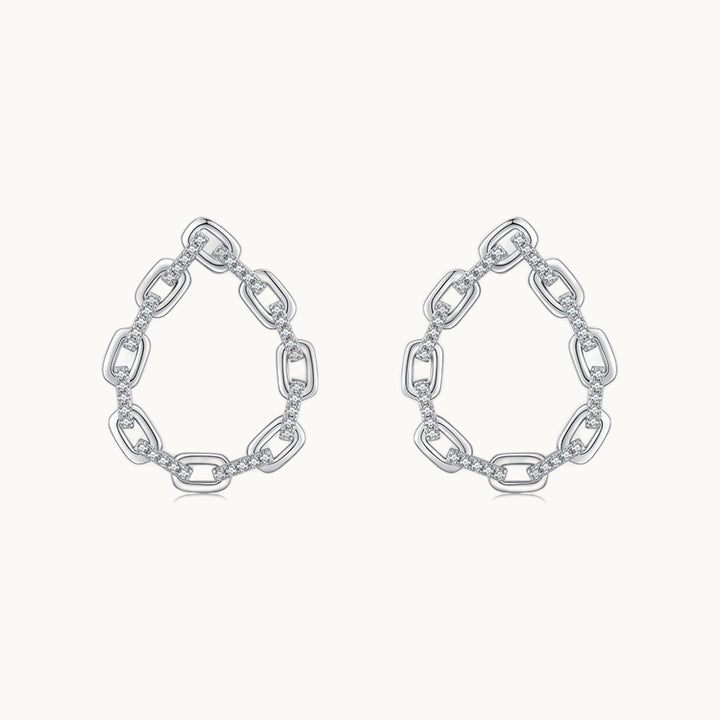 Une paire de boucles d'oreilles est exposée devant un fond beige.  Ce sont des boucles d'oreille en forme de goutte. Elles sont constituées de maillons de chaînes ornées de strass entre les maillons. Elles sont en argent 925. 