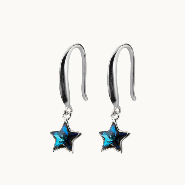 Une paire de boucles d'oreilles est exposée devant un fond beige.  Ce sont des boucles d'oreille en argent 925. La tige en argent pend et il y a au bout une étoile en diamant en cristal bleu.