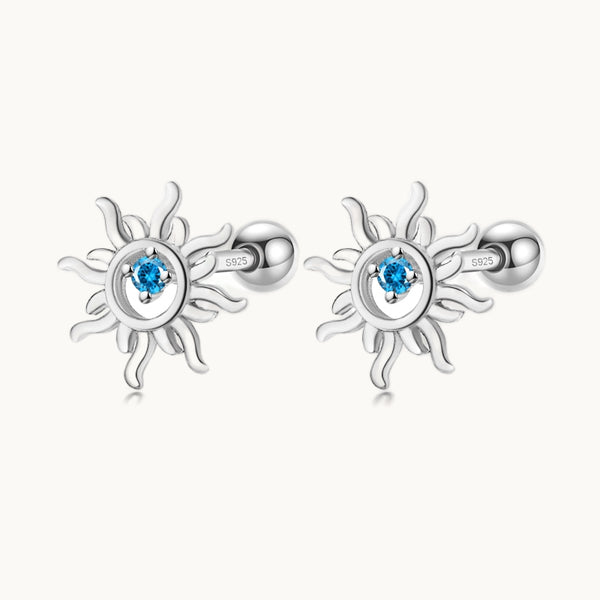 Une paire de boucles d'oreilles est exposée devant un fond beige.  Ce sont des soleil en argent avec un strass bleu. 