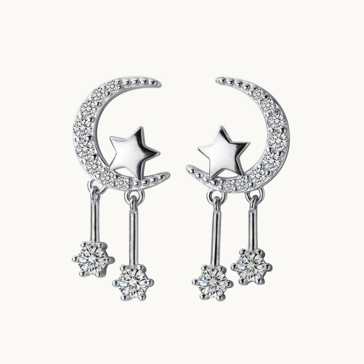 Une paire de boucles d'oreilles est exposée devant un fond beige.  Ce sont des boucles d'oreille en forme de lune. La lune est ornée de strass. Il y a une étoile accrochée à la lune. Sous la lune pendent deux tige sur lesquelles il y a un diamant de zirconium au bout. Elles sont en argent 925. 