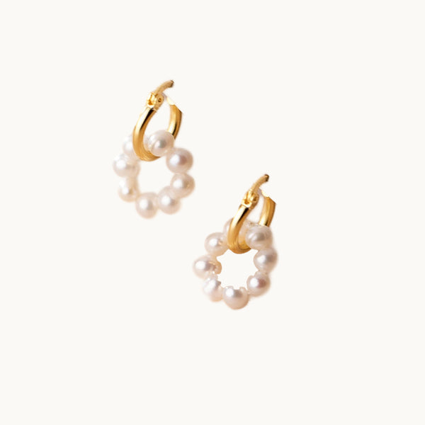 Une paire de boucles d'oreilles est exposée devant un fond beige.  Ce sont des doubles anneaux ; le premier est en or et le second en perles.