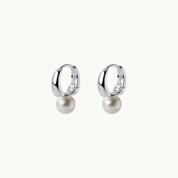 Une paire de boucles d'oreilles est exposée devant un fond beige.  Ce sont des anneaux épais sur lesquels est fixé une perle. Elles sont en argent 925.