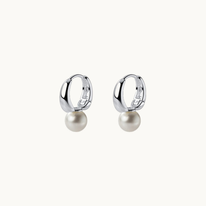 Une paire de boucles d'oreilles est exposée devant un fond beige.  Ce sont des anneaux épais sur lesquels est fixé une perle. Elles sont en argent 925.