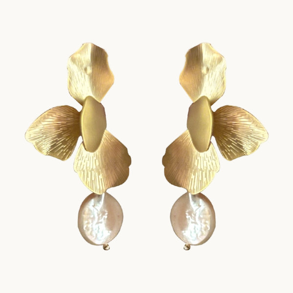 Une paire de boucles d'oreille est exposée devant un fond beige.  Ce sont des boucles d'oreille pendantes. Elles sont composées de pétales de fleurs. Il y a une perle plate ronde qui pend. Les pétales sont dorés. 