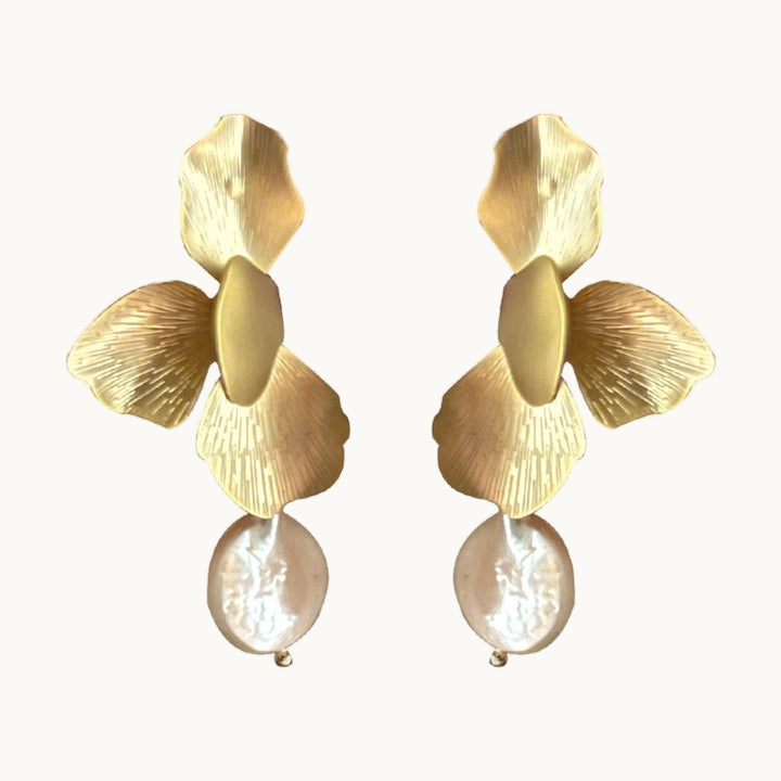 Une paire de boucles d'oreille est exposée devant un fond beige.  Ce sont des boucles d'oreille pendantes. Elles sont composées de pétales de fleurs. Il y a une perle plate ronde qui pend. Les pétales sont dorés. 