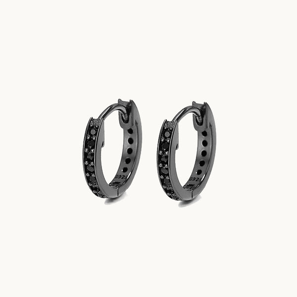 Une paire de boucles d'oreilles est exposée devant un fond beige.   Ce sont des anneaux ornés de diamants. Elles sont en argent 925 noir.