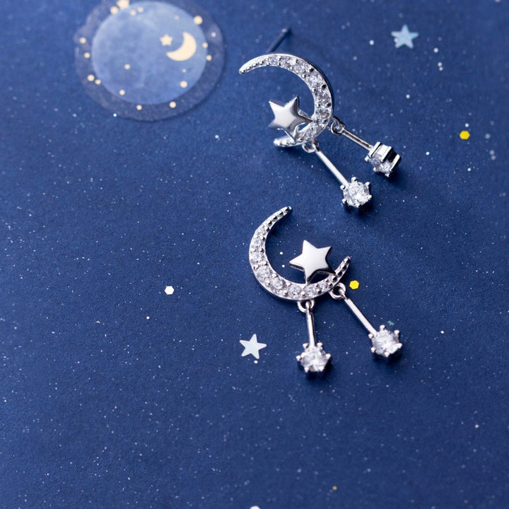 Une paire de boucles d'oreille en argent avec des diamants et des étoiles, évoquant un ciel étoilé. Les boucles d'oreille en forme de lune sont ornées de strass scintillants et d'une étoile. Des tiges pendantes avec des diamants de zirconium ajoutent une touche d'éclat et de mouvement. Parfait pour allier élégance intemporelle et modernité brillante.