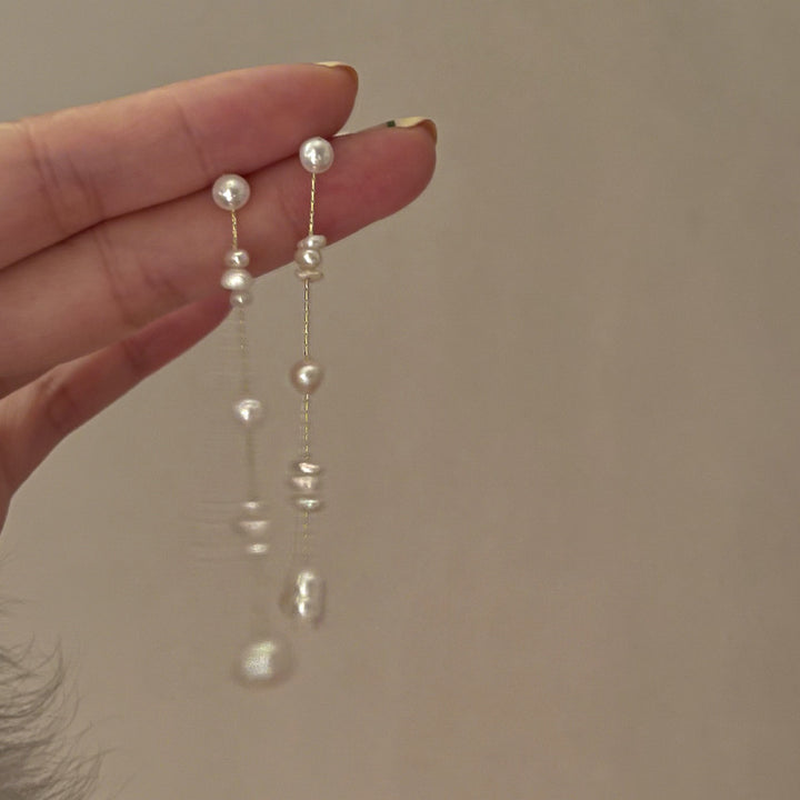 Une personne tenant une paire de boucles d'oreille longues et pendantes en perles d'eau douce et chaîne dorée. Un choix élégant pour toutes occasions.