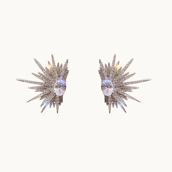 Une paire de boucles d'oreilles est exposée devant un fond beige.   Ce sont des boucles d'oreilles en diamants zirconium qui forment des demis soleils. 