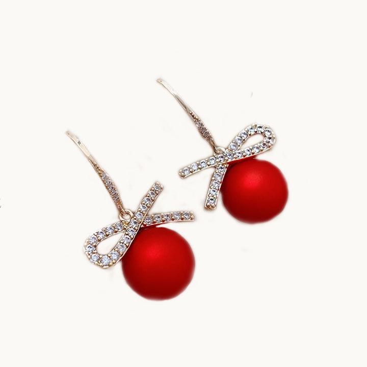 Une paire de boucles d'oreilles est exposée devant un fond beige.  Ce sont des boucles d'oreille pendante. Elles sont en forme de boule de noël rouge sur laquelle il y a un nœud en strass. Elles sont dorées. Elles sont idéales pour la période des fêtes de noël.