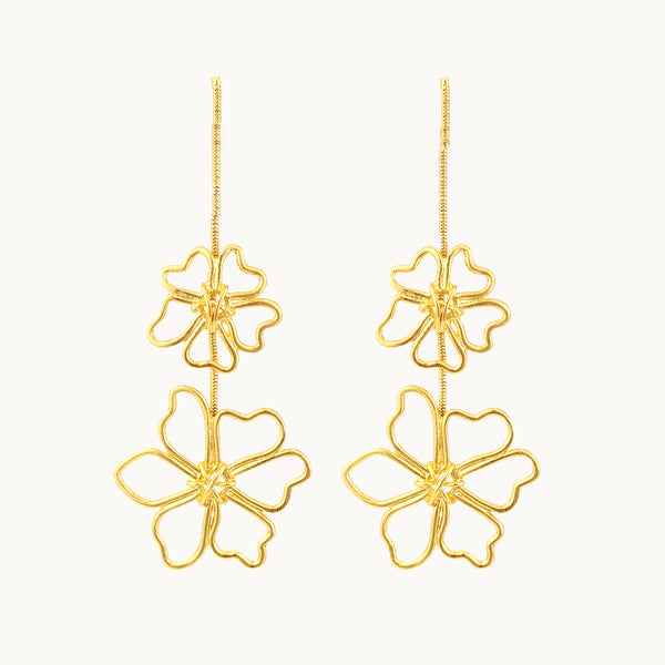 Une paire de boucles d'oreille est exposée devant un fond beige.  Ce sont des boucles d'oreille dorées longues et pendantes constituées d'une chaîne sur laquelle pend deux fleurs métalliques. Il y a une petite fleur et en dessous une grande fleur. 