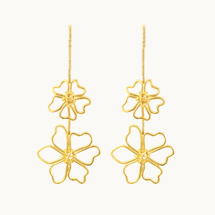 Une paire de boucles d'oreille est exposée devant un fond beige.  Ce sont des boucles d'oreille dorées longues et pendantes constituées d'une chaîne sur laquelle pend deux fleurs métalliques. Il y a une petite fleur et en dessous une grande fleur. 