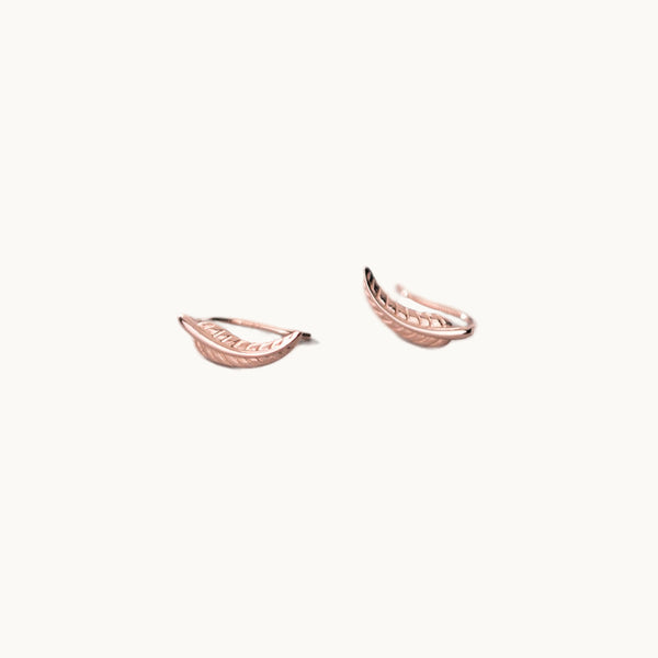 Une paire de boucles d'oreilles est exposée devant un fond beige.  Ce sont des boucles grimpantes en forme de plume. Ce sont des boucles or rose. 