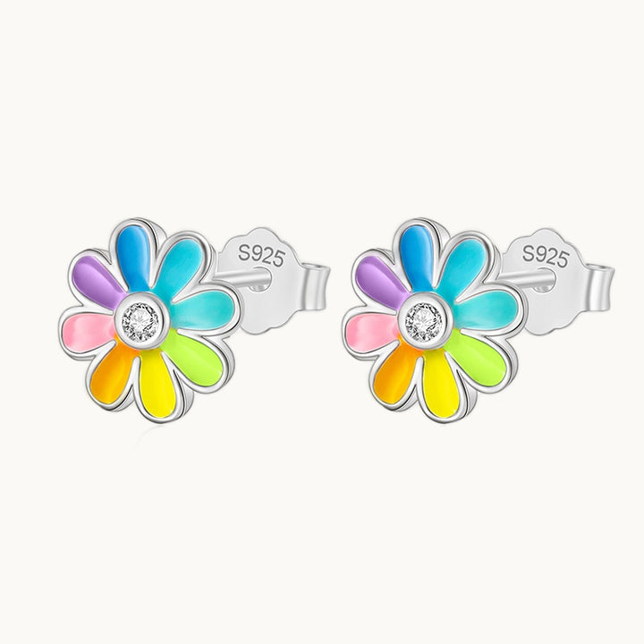 Une paire de boucles d'oreilles est exposée devant un fond beige.  Ce sont des boucles en forme de marguerite avec les pétales colorés et un strass au coeur de la fleur. Elles sont en argent 925 et les pétales sont multicolores. 