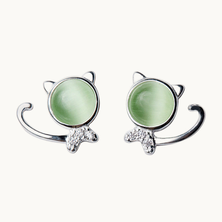 Une paire de boucles d'oreilles est exposée devant un fond beige.  Ce sont des boucles d'oreille en forme de chat. La tête est ornée d'un cristal vert clair et le corps du chat est en strass de zirconium.  Elles sont en argent 925. 