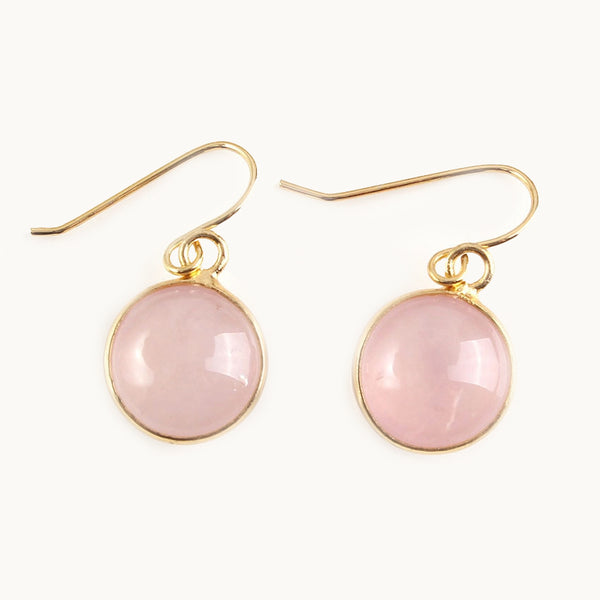 Une paire de boucles d'oreilles est exposée devant un fond beige.  Ce sont des boucles d'oreille dorées pendantes. Elles ont au bout une pierre naturelle ronde en quartz rose. 