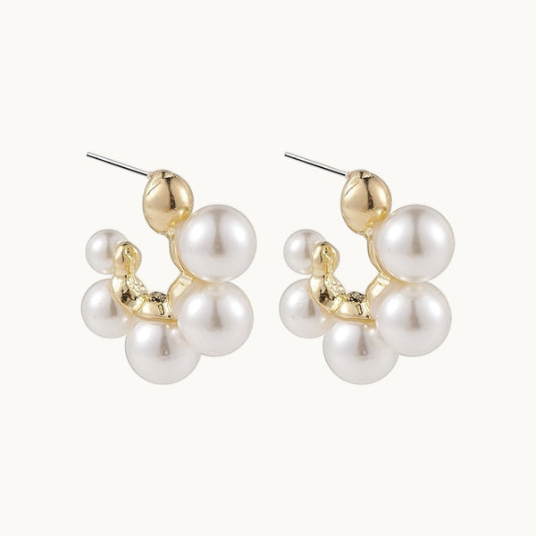 Une paire de boucles d'oreilles est exposée devant un fond beige.  Ce sont des créoles dorées qui ne font pas le tour de l'oreille, elles ne sont pas fermées. Elles sont ornées de très grosses perles. 