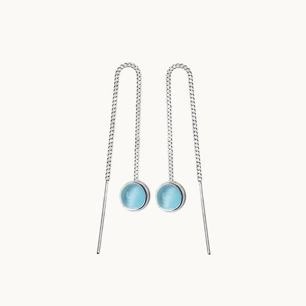 Une paire de boucles d'oreilles est exposée devant un fond beige.  Ce sont des boucles d'oreilles pendantes. Elles sont en chaine avec une pierre ronde opale bleue au bout. Elles sont en argent 925. 